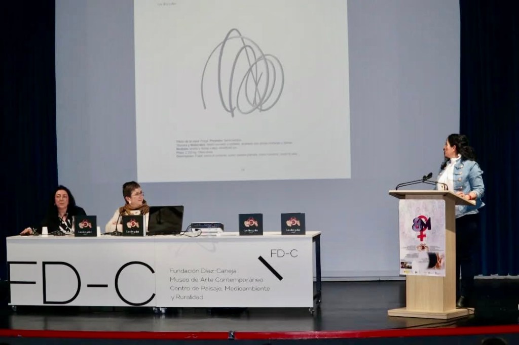 Acto de presentación del libro en la Fundación Díaz-Caneja. Dos mujeres en la mesa presidencial con ejemplares del libro. Al atril, una artista presenta; en la pantalla, líneas abstractas entrelazadas.