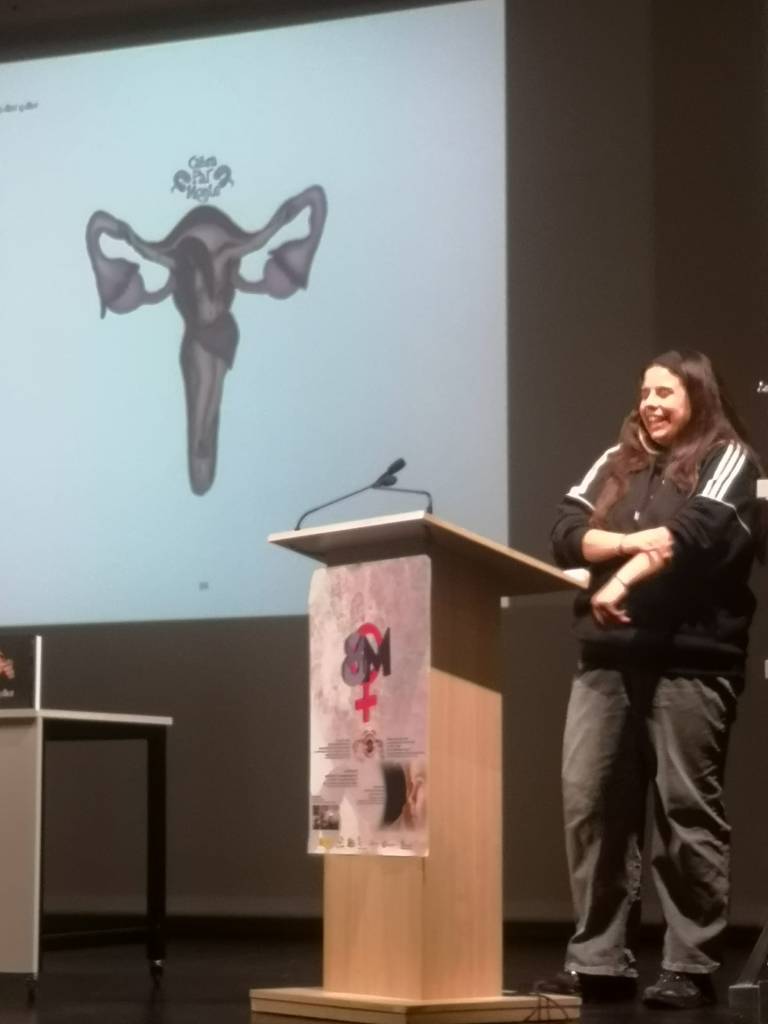 Presentación del libro. Una artista habla en el atril. En la pantalla, ilustración de órganos reproductores femeninos en tonos oscuros sobre fondo azul, obra de Cabra pal Monte.