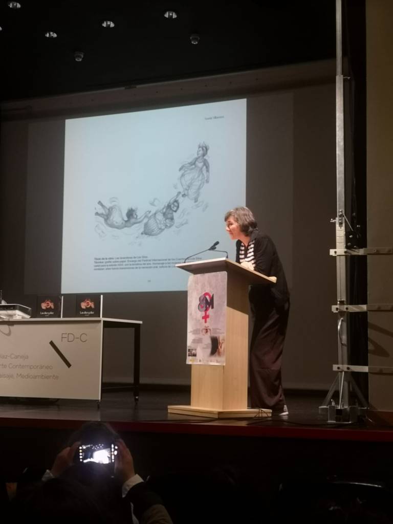Presentación del libro. Una artista habla en el atril. En la pantalla, ilustración a grafito de figuras femeninas en movimiento.