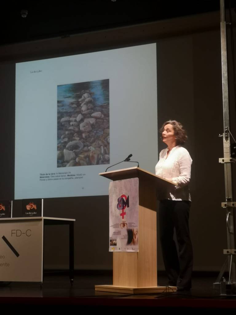 Presentación del libro. Una artista habla en el atril. En la pantalla, obra pictórica con detalle de guijarros y agua.