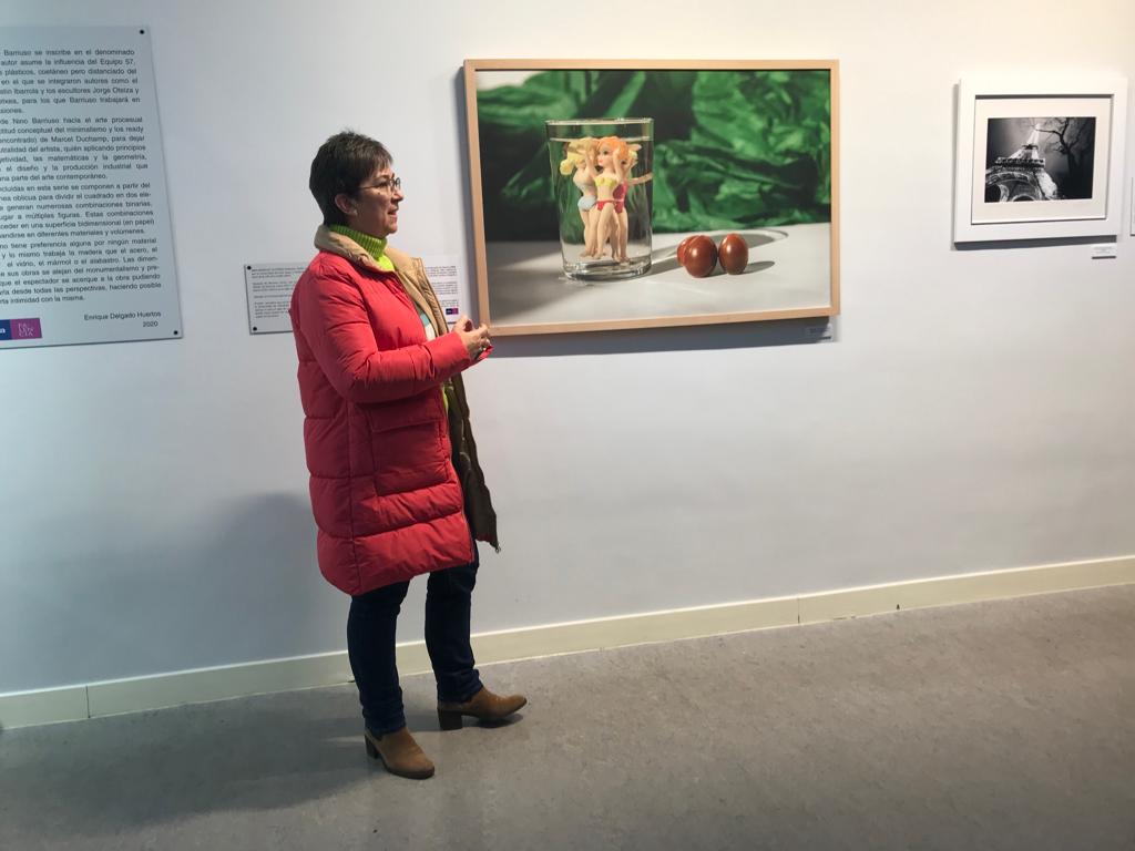 Mujer con abrigo rojo y gafas habla junto a la fotografía del vaso colgada en la pared.