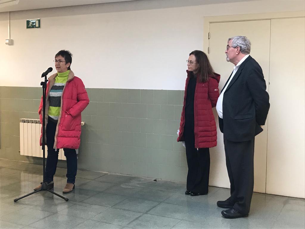 Mujer con abrigo rojo habla ante un micrófono de pie. A la derecha la escuchan dos personas, una mujer con abrigo rojo y un hombre con traje oscuro.