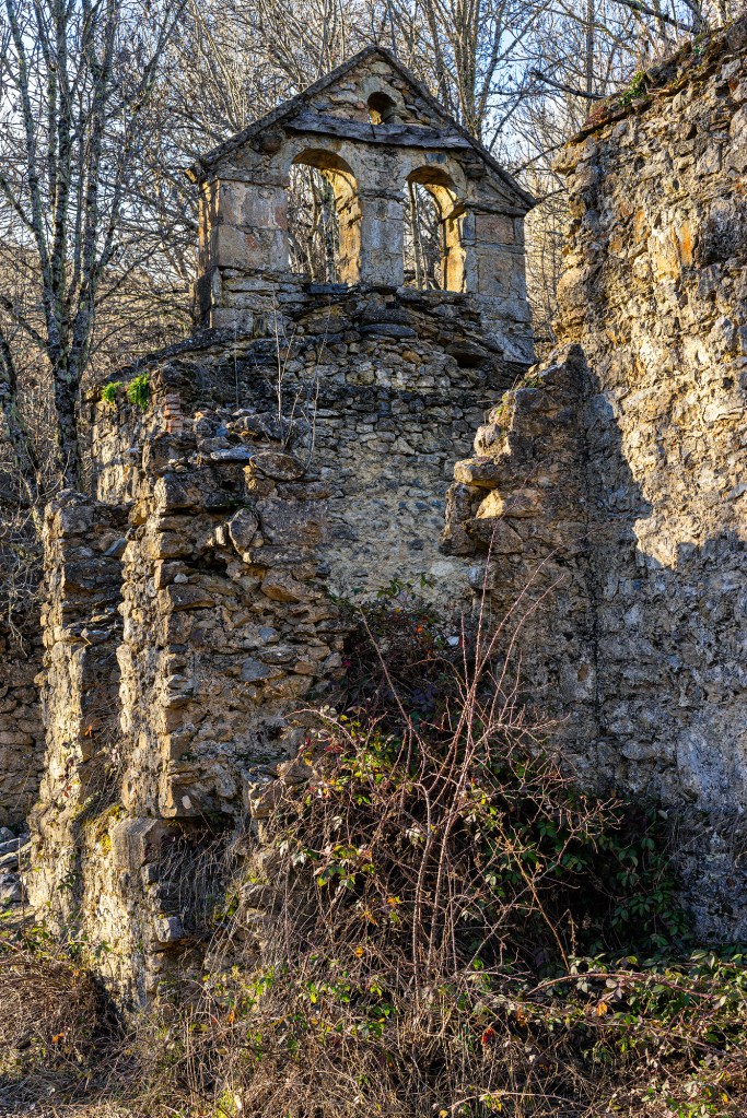 Ruina de la iglesia de Valsurbio: espadaña de piedra con dos arcos y un óculo sobre muros derruidos, invadida por zarzas, con árboles desnudos y cielo azul al fondo.